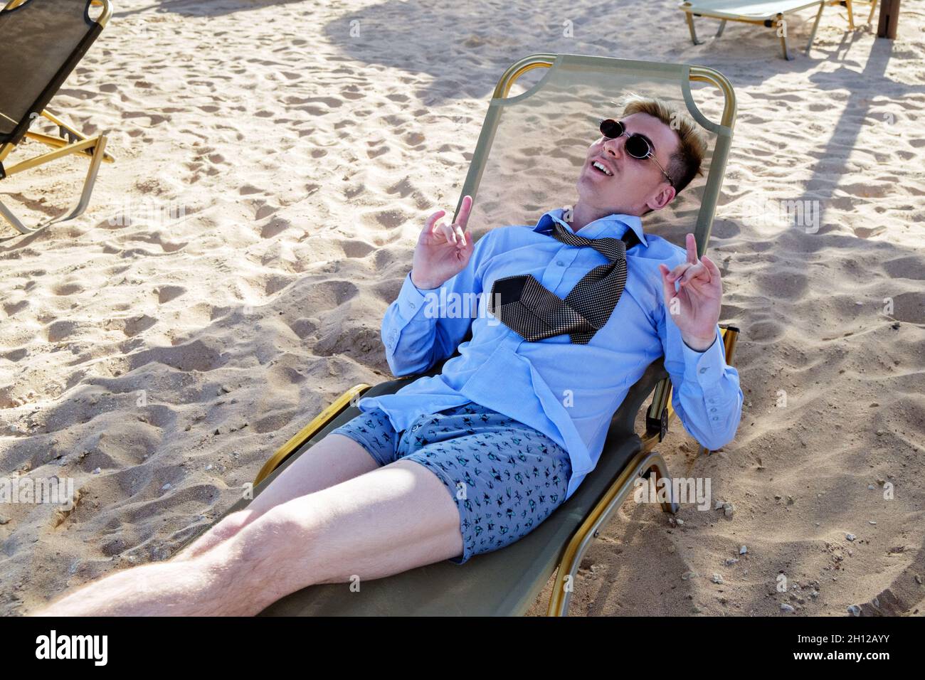 Il giovane uomo si trova su un lettino al centro della spiaggia in una maglietta, cravatta e pantaloncini da boxer. Mostra gli avvisatori acustici manuali. Gode di riposo Foto Stock