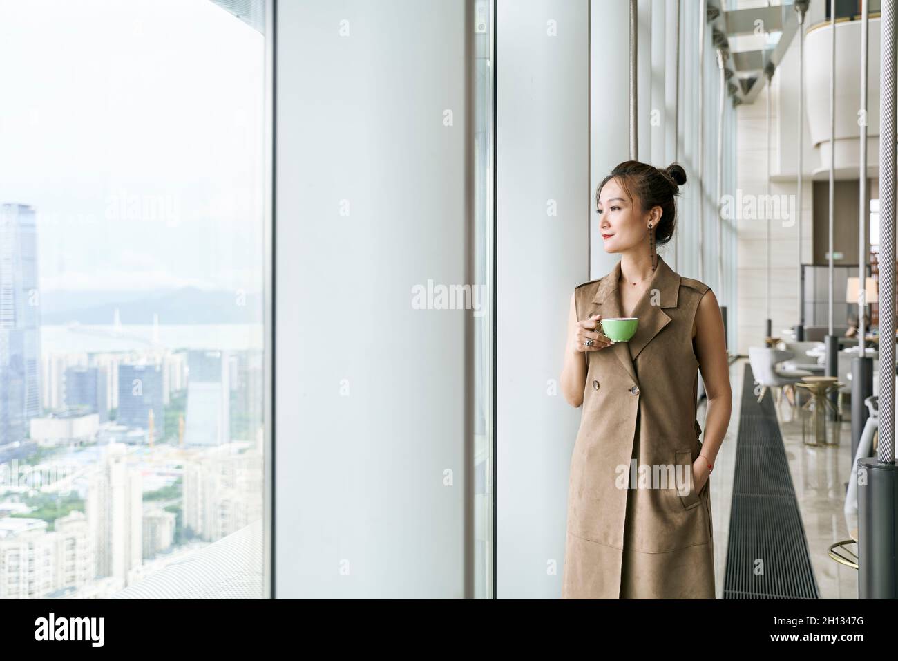 donna asiatica matura in piedi dalla finestra che guarda attraverso la finestra che tiene una tazza di caffè o tè Foto Stock