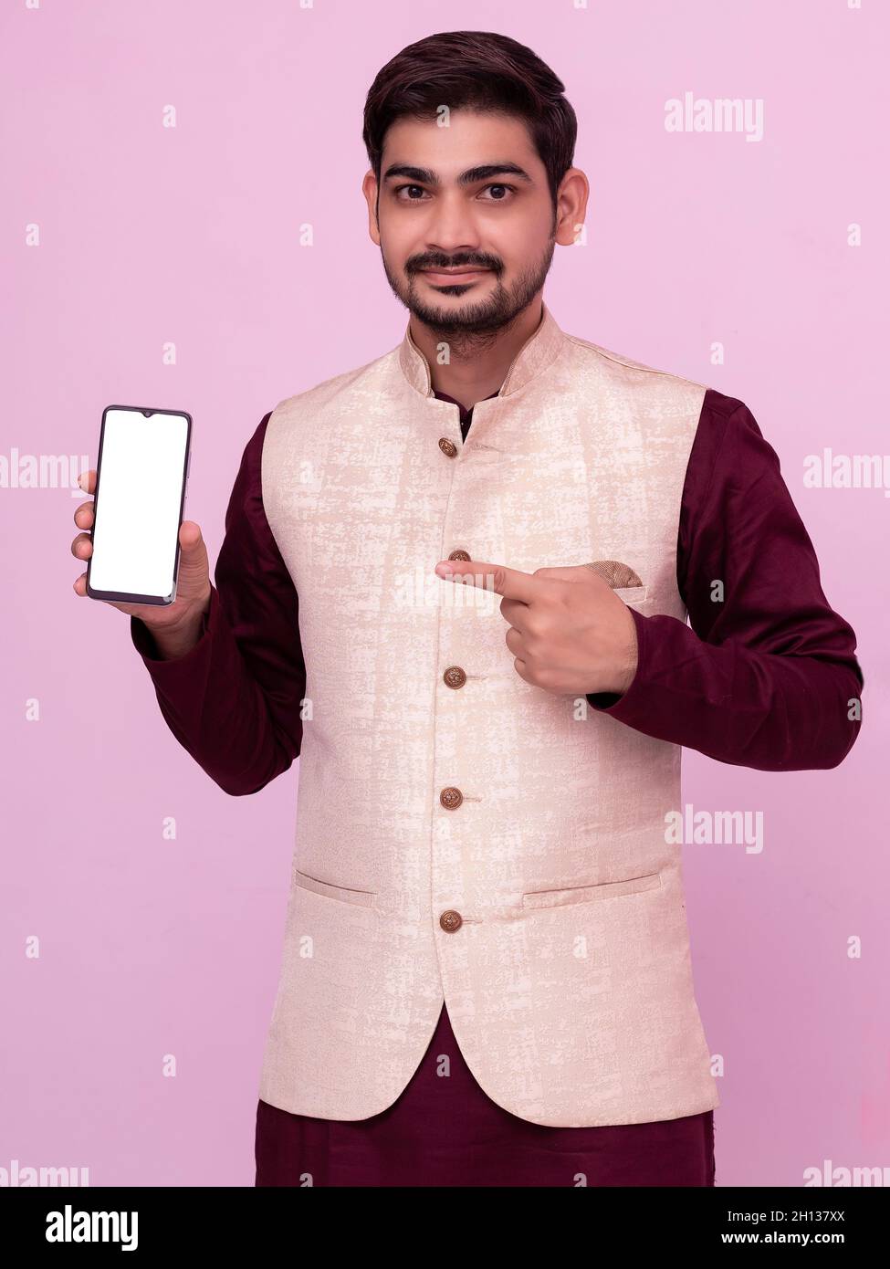 Ritratto di uomo con barba in costume tradizionale puntando al telefono cellulare e sorridendo alla macchina fotografica. Giovane uomo indiano in abbigliamento tradizionale e mostrando smartph Foto Stock
