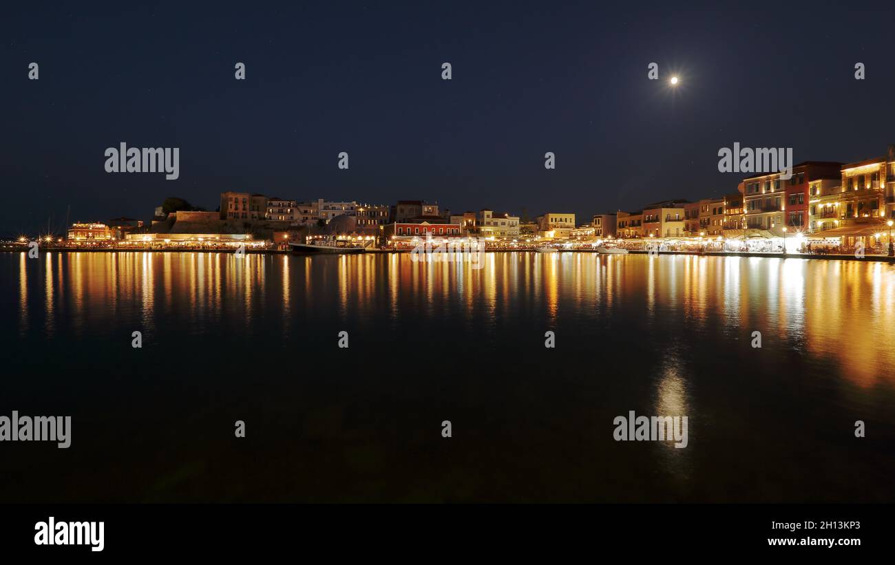 La Canea di notte, la baia del mare, gli edifici illuminati si riflettono nella superficie dell'acqua, la luna piena nel cielo Foto Stock