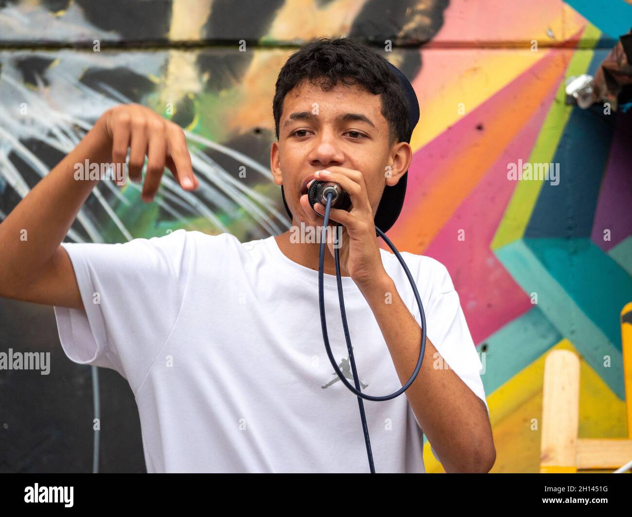 Medellin, Antioquia, Colombia - 6 gennaio 2021: Giovane uomo latino con capelli ricci canta Hip-Hop con microfono nero contro Muro colorato Foto Stock