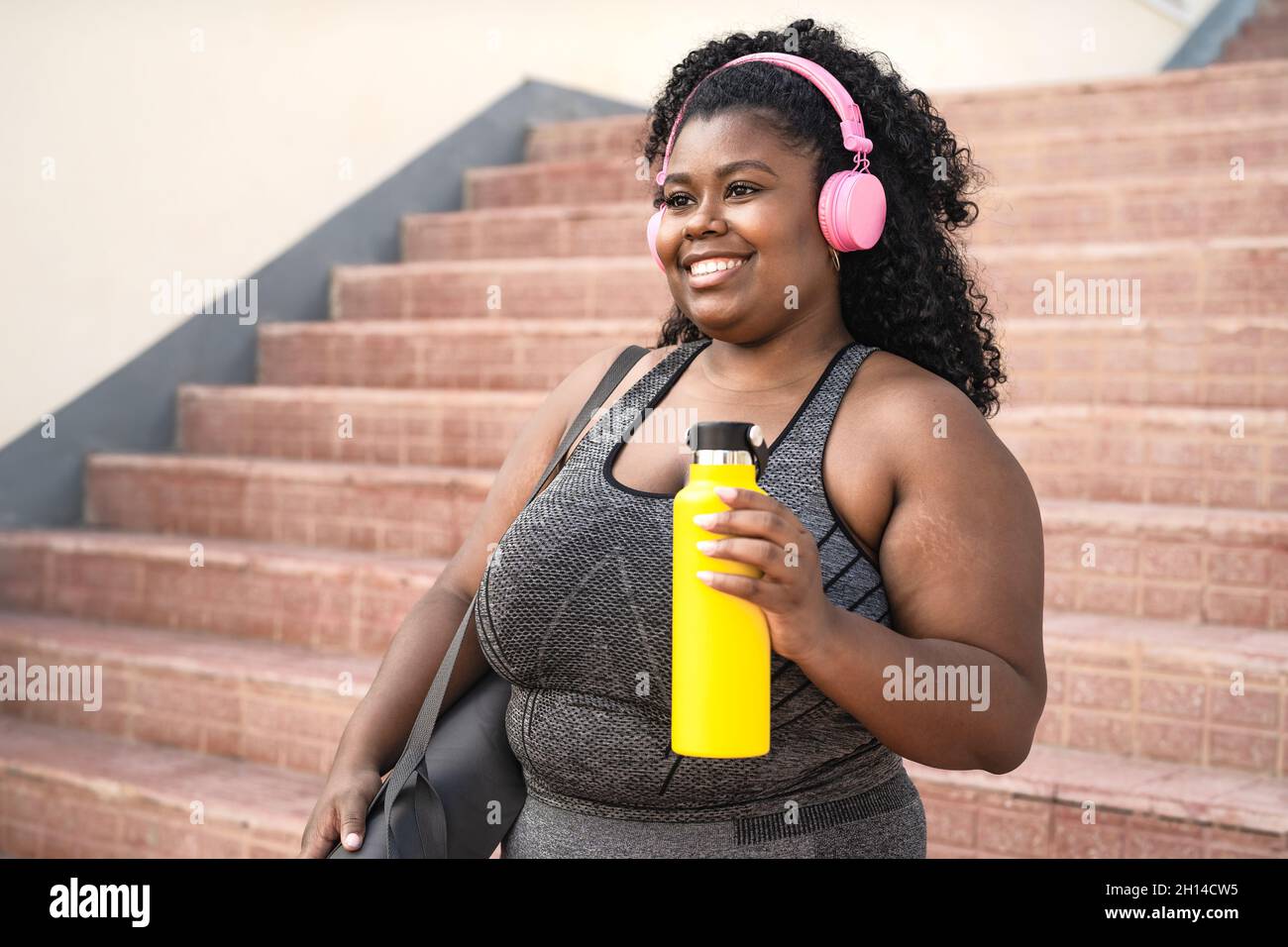 Una donna africana di dimensioni più felici che fa la routine di allenamento mentre ascolta la musica con le cuffie wireless all'aperto Foto Stock