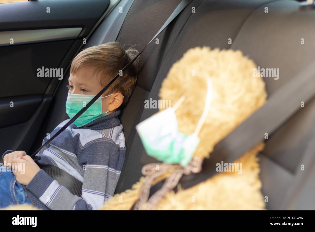 Un ragazzo carino di sette anni che indossa una maschera medica si siede in un'auto in una calda giornata di sole autunno durante la pandemia di coronavirus. Nelle vicinanze si trova un orso giocattolo. Selez Foto Stock