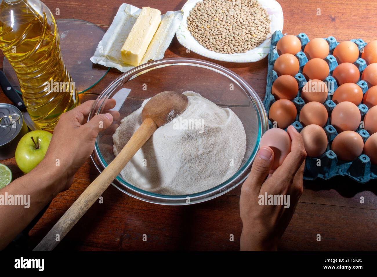 Le mani tagliano il limone con il coltello sul pannello di legno. Concetto di cucina domestica. Contenitore di farina, uova, burro e ingredienti da cucina su tavola di legno. Foto Stock