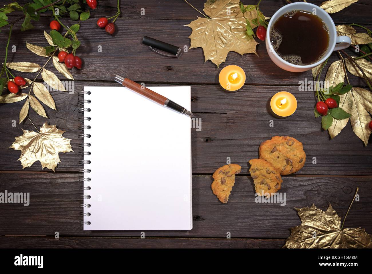 pagina vuota di un notebook a spirale mock in su su su un tavolo di legno rustico scuro con caffè, foglie d'autunno dorate, candele e biscotti per una vacanza stagionale a. Foto Stock
