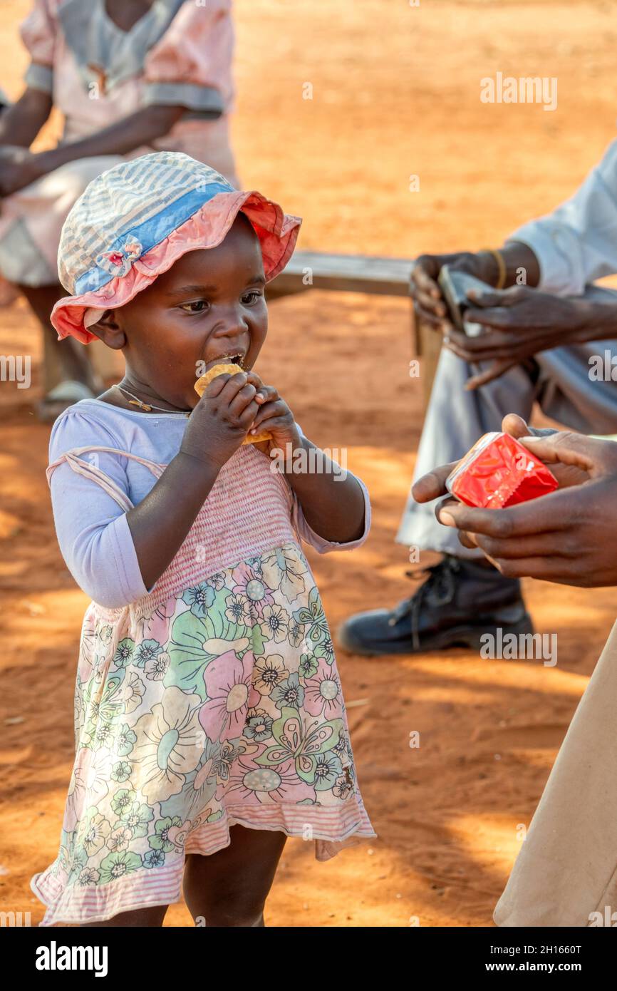 Ragazza africana in un villaggio in Botswana seduta di fronte alla capanna nella sporcizia, giorno di sole, con la famiglia che mangia biscotti Foto Stock