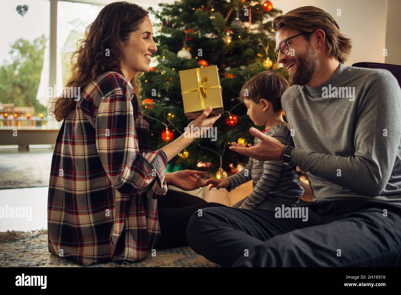 Coppia seduta da albero di Natale con il loro figlio che si scambiano i regali a casa. Bella piccola famiglia che festeggia il Natale a casa. Foto Stock