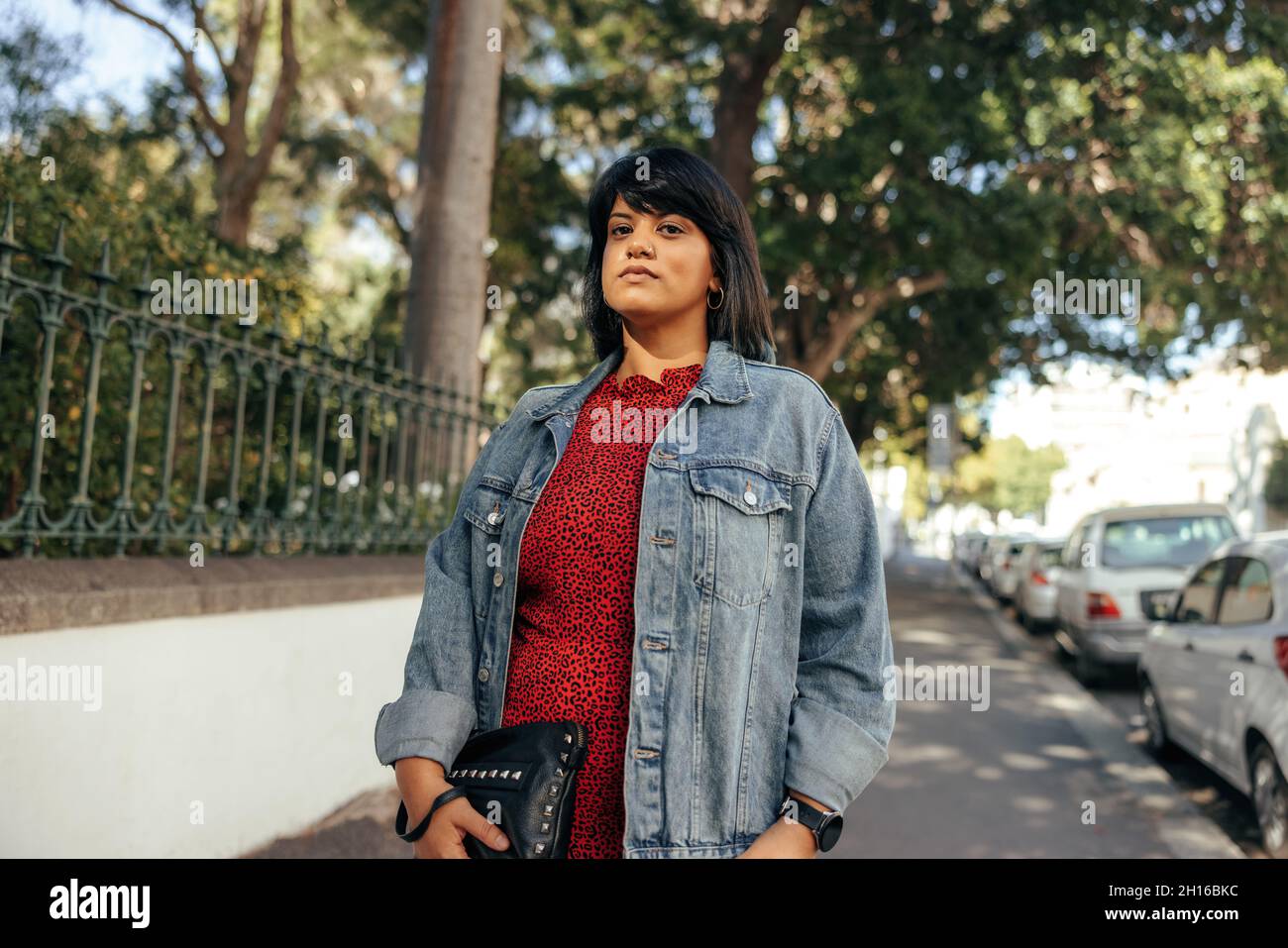 Donna di metà età adulta in piedi su un marciapiede in città. Donna sicura che guarda la telecamera mentre si trova accanto a un parco. Donna che indossa un denim ja Foto Stock