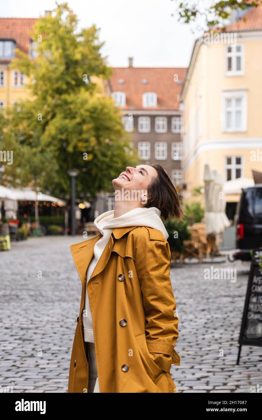 Affascinante donna in giallo cappotto e felpa con cappuccio in piedi sul  marciapiede sulla strada mentre sorride e guarda in su in autunno Foto  stock - Alamy