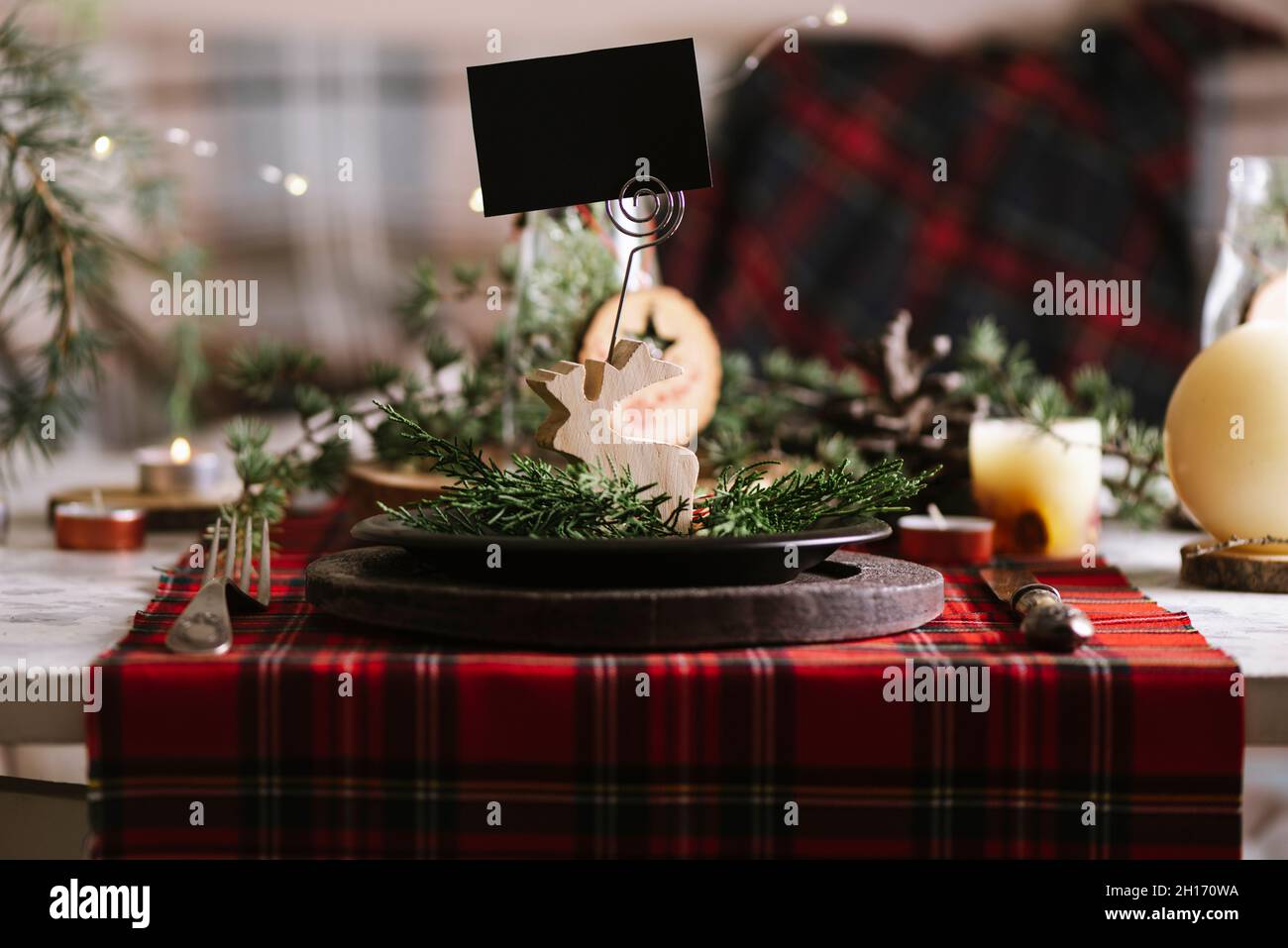 Tavolo di Natale con renna di legno con carta del nome vuota su piastra di ceramica su tovaglia a scacchi rosso sullo sfondo Foto Stock
