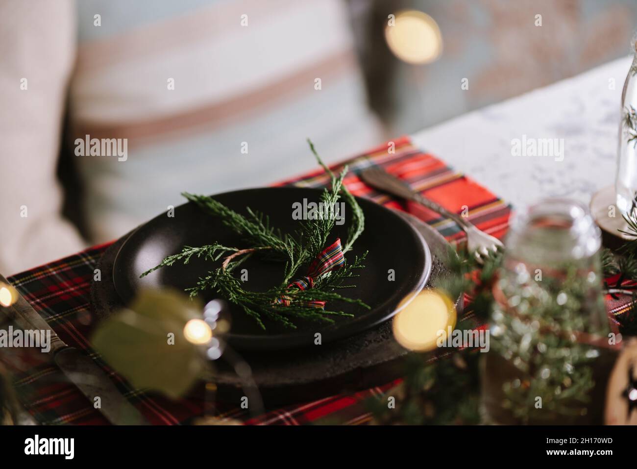 Tavolo di Natale con corona su piastra di ceramica su tovaglia a scacchi rossi sullo sfondo con luci Foto Stock
