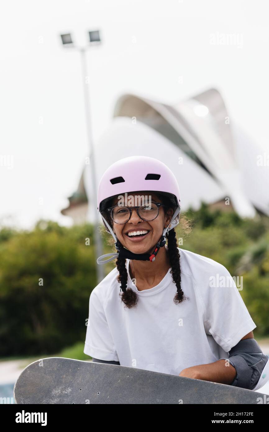 Sorridente giovane afroamericana in occhiali con casco protettivo lilla chiaro e gommini grigio pattinaggio nel parco Foto Stock