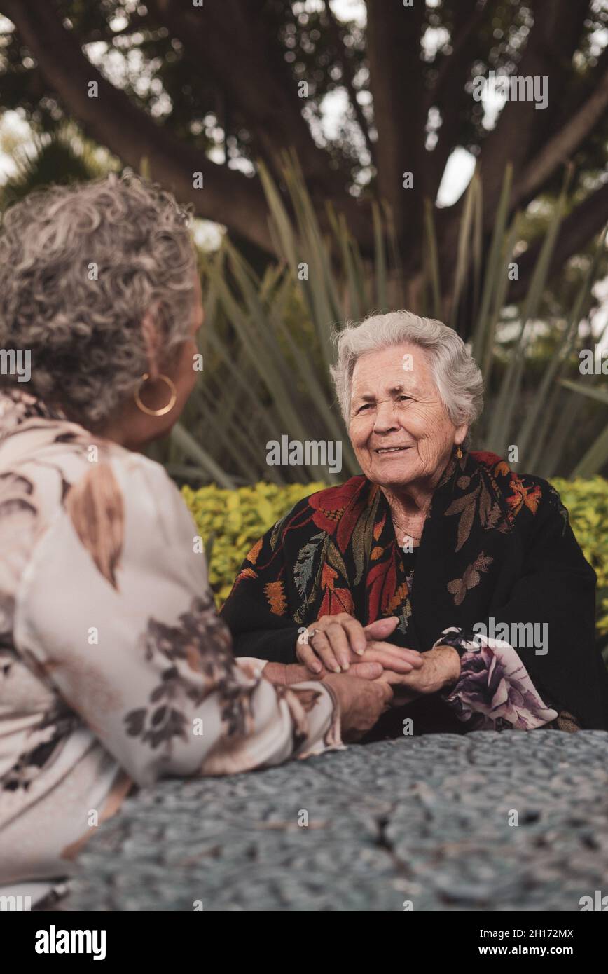 Vecchia femmina sorridente e tenuta le mani mentre si siede a tavola in un cortile esotico con palme e cespugli e godendo di conversazione con la donna anziana ven Foto Stock