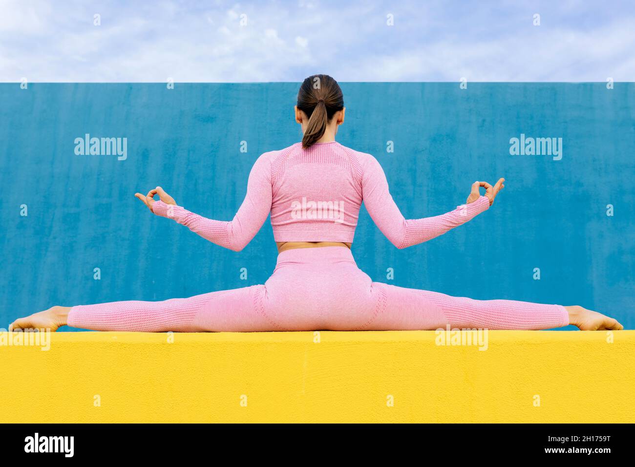 Vista posteriore di donna snella seduta in Split facendo gyan mudra mentre si siede su un alto muro giallo su sfondo blu Foto Stock