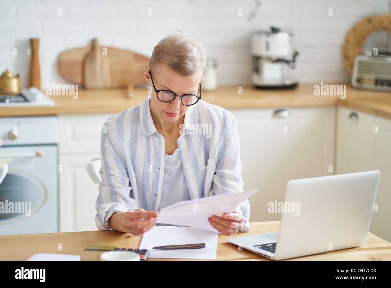 Donna anziana focalizzata che indossa occhiali che lavorano con documenti mentre si siede nella cucina moderna Foto Stock
