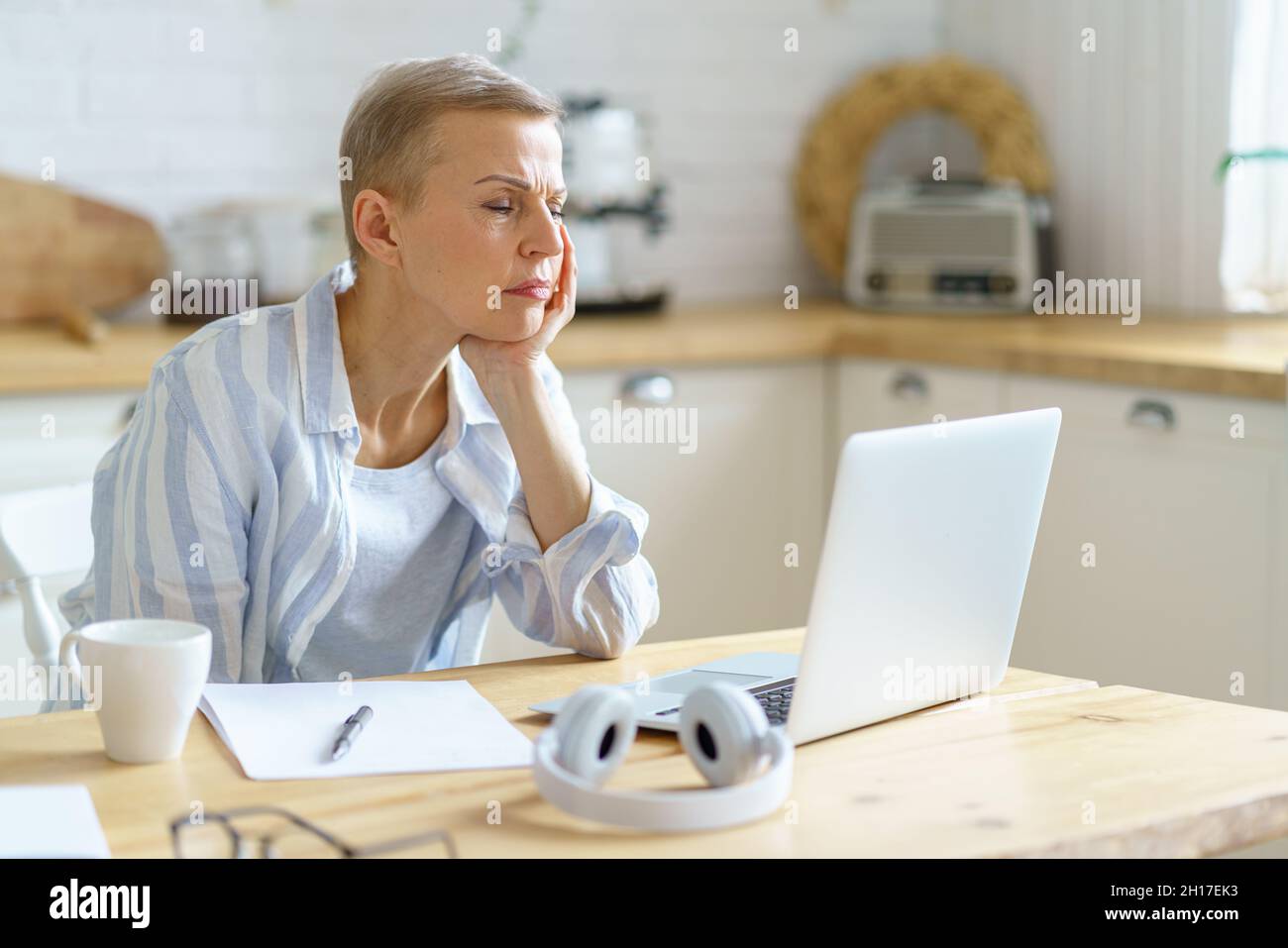 Donna anziana confusa che tiene la testa nelle mani e guarda sul laptop mentre lavora online da casa Foto Stock