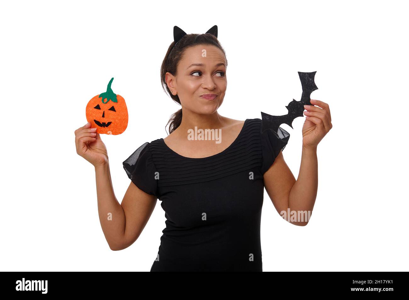Sorprende misteriosa sorridente donna africana in cerchio con orecchie di gatto, vestita di nero, tiene la zucca e il pipistrello fatti a mano di arancia, si pone contro il muso Foto Stock
