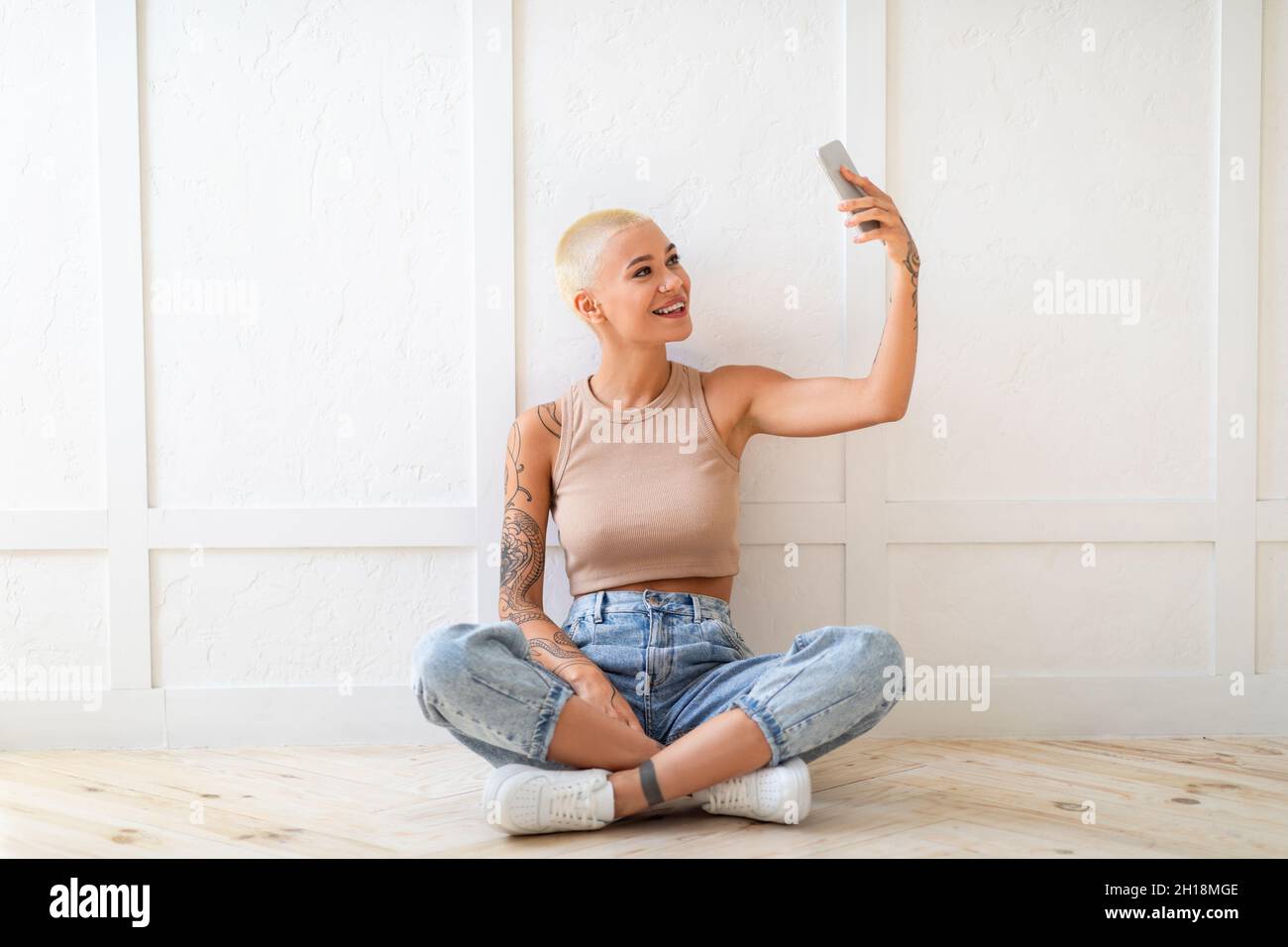 Giovane donna elegante che porta selfie su smartphone per social media o amici, seduto sul pavimento sopra una parete bianca Foto Stock