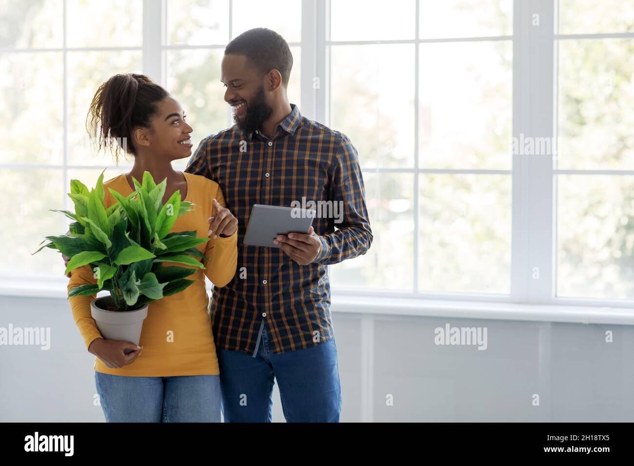 Allegro uomo e donna afroamericana millenario guardare l'un l'altro tenere tablet e pianta sullo sfondo finestra Foto Stock