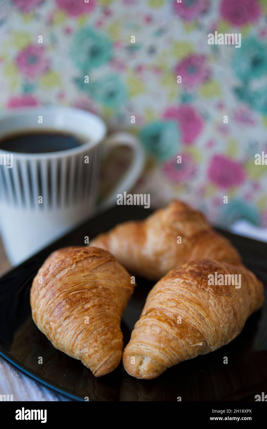 Spuntino con tre cornetti alla burro e fiammanti su un piatto nero e un caffè nero in una tazza grande, colazione continentale semplice e fresca Foto Stock