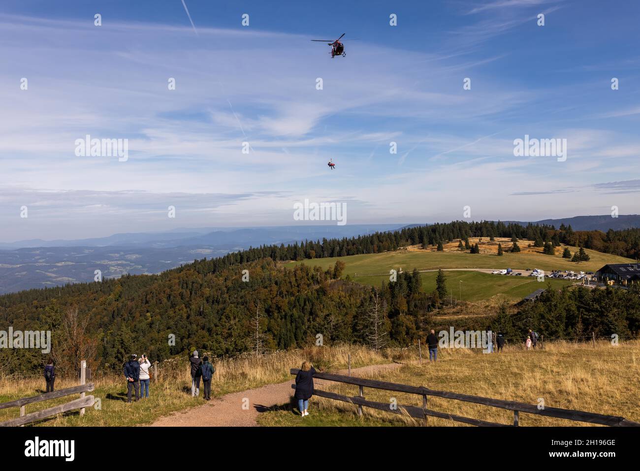 Waldkirch, Germania. 15 ottobre 2021. Gli spettatori si levano in piedi sulla cima del Kandel mentre sullo sfondo due persone appendono su una corda sotto un elicottero di salvataggio del Luftrettung DRF. Durante un allenamento con verricello, l'equipaggio della stazione di Friburgo della DRF Luftrettung (cartello di chiamata Christoph 54) si pratica con il verricello sotto un elicottero insieme al Bergwacht Schwarzwald. Questo verricello può essere utilizzato per salvare i pazienti da terreni impraticabili, nonché per trasportare medici e paramedici in regioni difficili da raggiungere. Credit: Rin/dpa/Alamy Live News Foto Stock