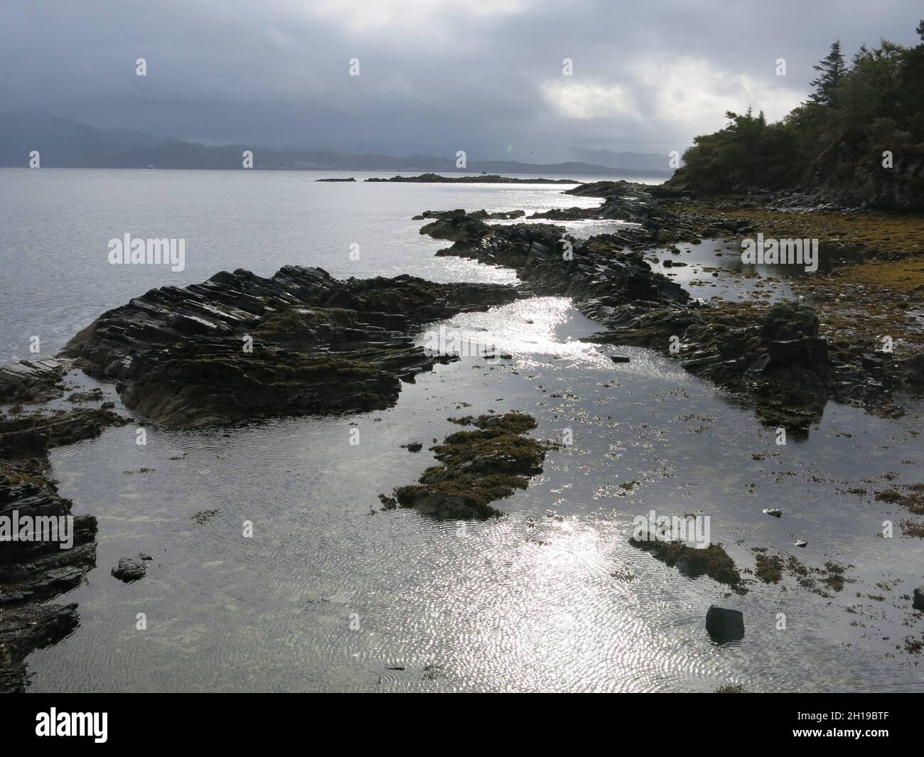 Paesaggio costiero sulla penisola di Sleat, Isola di Skye: Luce solare applaiata sull'acqua e una costa rocciosa con lontane colline grigie all'orizzonte Foto Stock