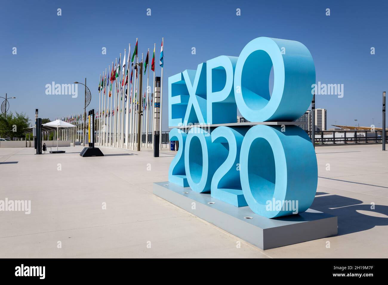 Dubai, Emirati Arabi Uniti, 17.10.2021. Expo 2020 cartello con il logo blu all'ingresso del sito dell'Expo Mobility District con bandiere nazionali dei partecipanti sullo sfondo. Foto Stock