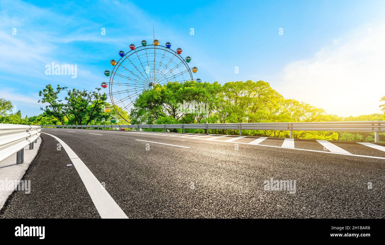 Strada asfaltata vuota e ruota di ferro con foresta verde sotto il cielo blu. Foto Stock