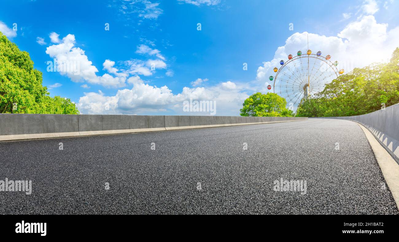 Strada asfaltata vuota e ruota di ferro con foresta verde sotto il cielo blu. Foto Stock