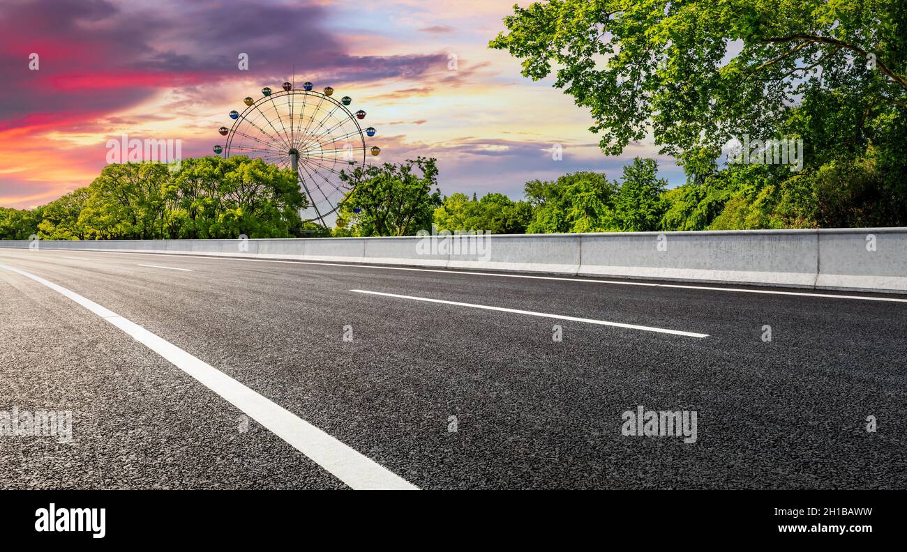 Strada asfaltata vuota e ruota di ferro con foresta verde al tramonto. Foto Stock