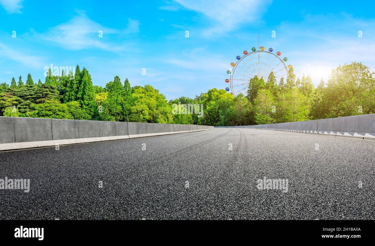 Strada asfaltata vuota e ruota di ferro con foresta verde sotto il cielo blu. Foto Stock