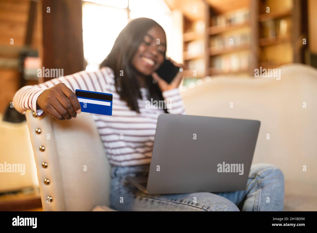 Shopping online. Signora nera millennial con il laptop usando la carta di credito, avendo chiamata telefonica per ordinare le merci sul fotoricettore da casa Foto Stock