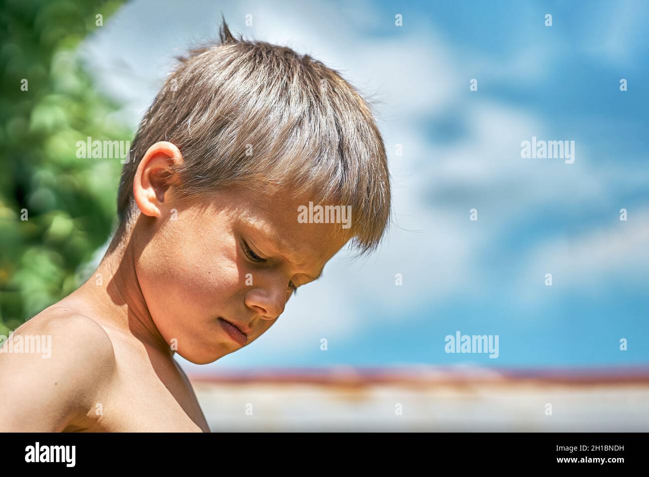 Scolboy junior arrabbiato con capelli biondi ruvidi in posa per la macchina fotografica nel cortile della casa d'estate su bella vista giorno d'estate vicino Foto Stock