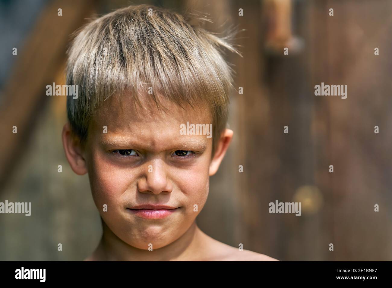 Scolboy junior arrabbiato con capelli biondi ruvidi in posa per la macchina fotografica nel cortile della casa d'estate su bella vista giorno d'estate vicino Foto Stock