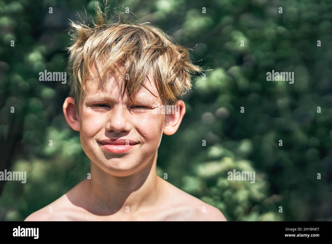 Sorridente scolaro junior con capelli biondi diseveled ruvidi in posa per la macchina fotografica nel parco verde in bella vista giorno d'estate vicino Foto Stock