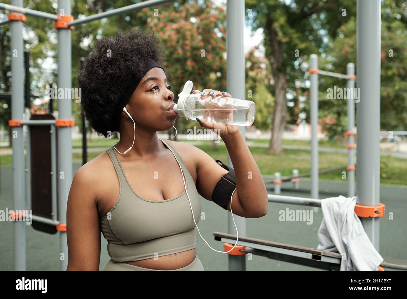 Giovane donna assetata in tuta acqua potabile da bottiglia di plastica contro impianti sportivi Foto Stock