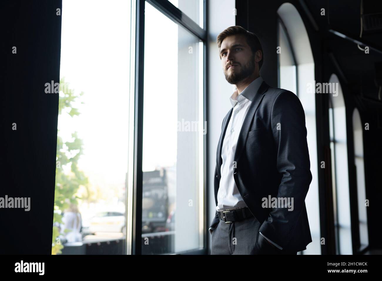 Uomo d'affari giovane sicuro che guarda la macchina fotografica mentre si trova vicino alla finestra di un edificio di uffici. Foto Stock