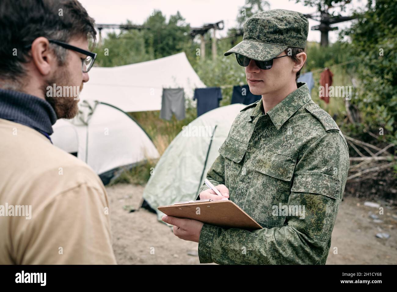 Giovane uomo in uniforme verde che guarda il documento nelle sue mani durante l'interazione con il volontario Foto Stock