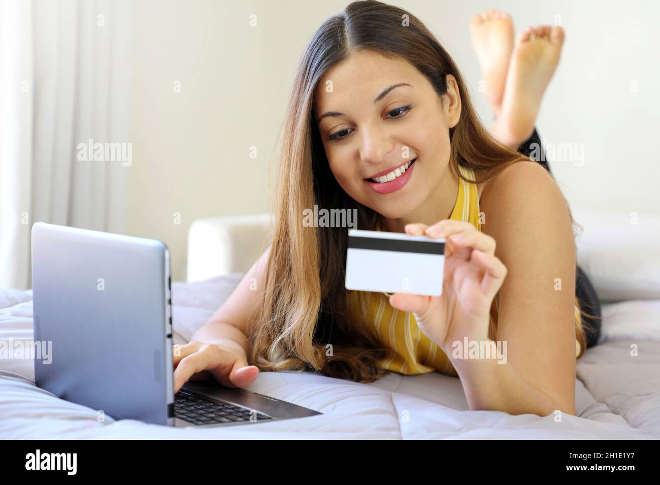 Sorridente giovane donna rilassata sdraiata sul letto leggendo il suo numero di carta di credito e inserire il numero sul notebook facendo shopping online confortevole a casa Foto Stock