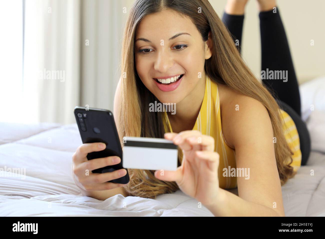 Felice giovane donna rilassata sdraiata sul letto leggendo il suo numero di carta di credito e inserire il numero sullo smartphone facendo shopping online confortevole a casa. E Foto Stock