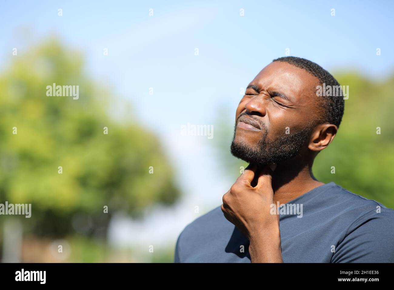 Uomo con pelle nera che soffre mal di gola in piedi in un parco una giornata di sole Foto Stock