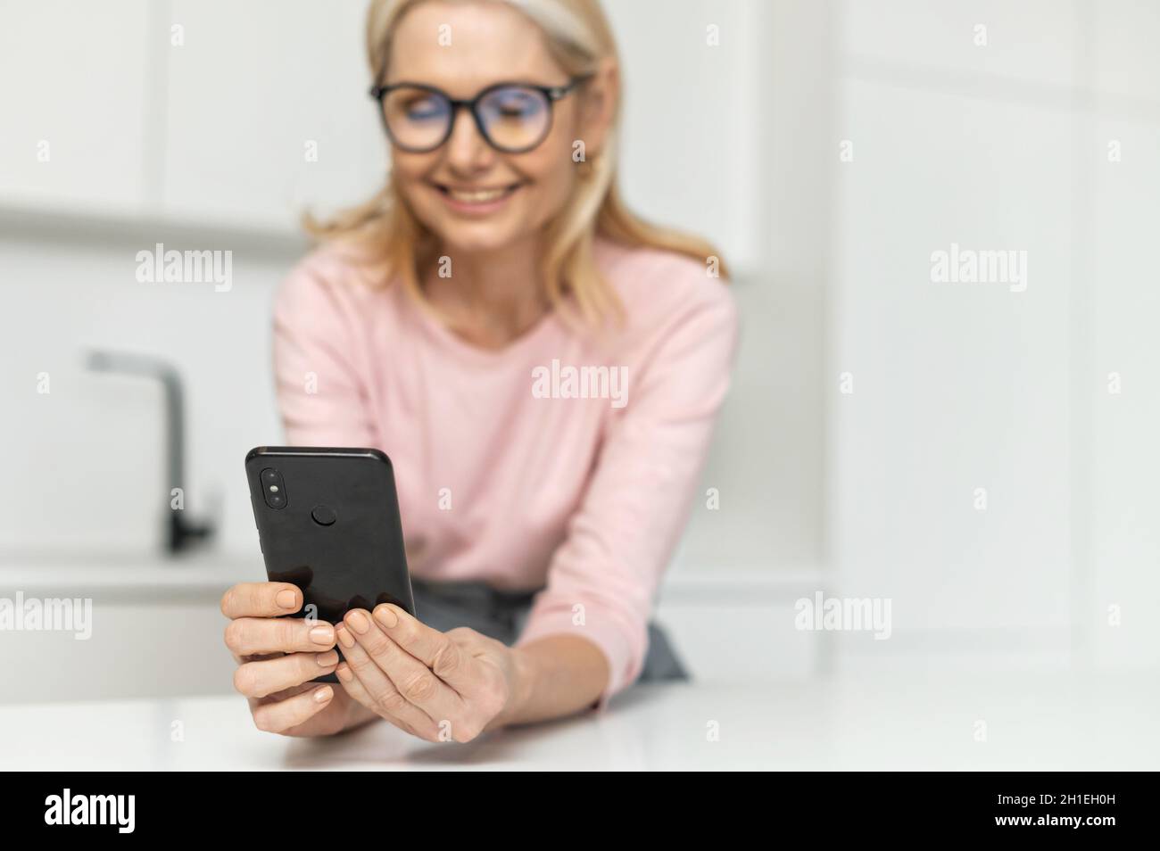 Sorridente ragazza bionda affascinante trascorre il tempo libero con uno smartphone a casa, controllare le e-mail, shopping online, scorrimento news feed, sorridente mezza età sesso femminile web surf, fuoco selettivo al telefono Foto Stock