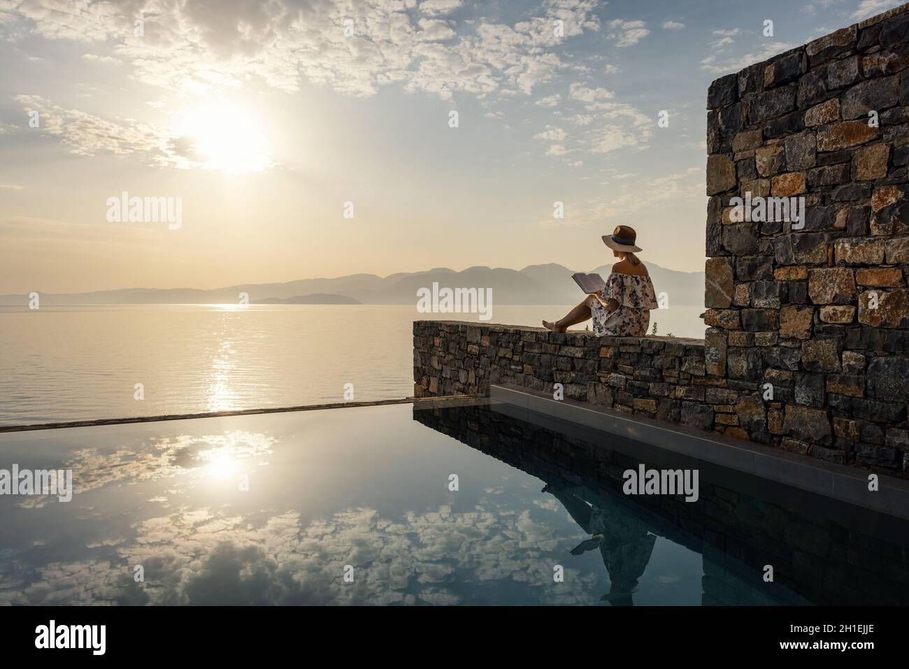 concetto di relax e meditazione - donna che legge un libro vicino alla piscina con vista sul mare all'alba in hotel di lusso Foto Stock