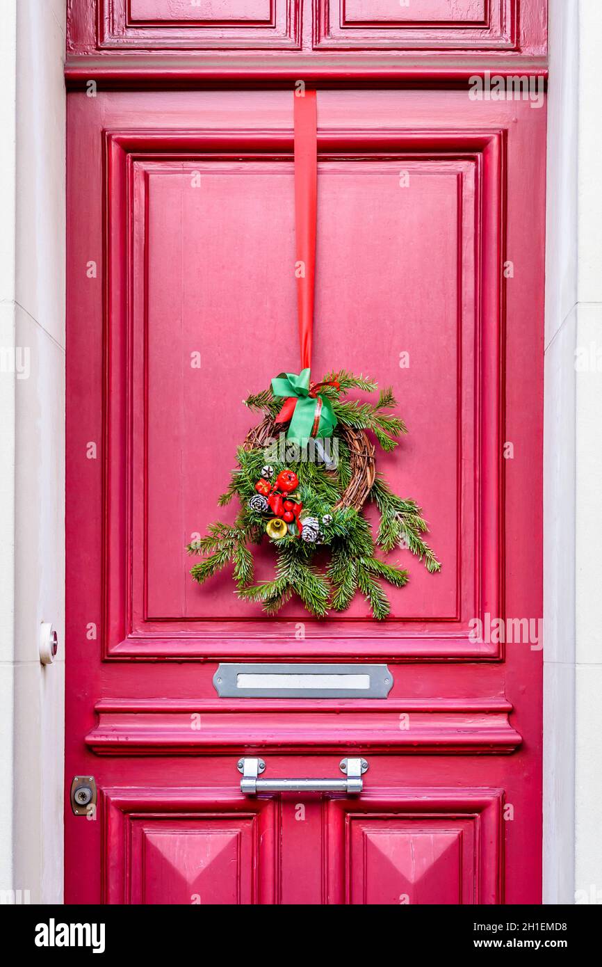 Vista frontale di una corona di Natale fatta di rami di abete, coni di pino e nastri, appesi sulla porta rossa con modanature di una casa cittadina. Foto Stock