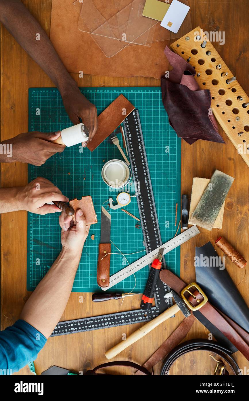 Vista dall'alto sfondo di due artigiani irriconoscibili che fanno pezzo di pelle fatto a mano su tavolo di legno in laboratorio di pelletteria, spazio copia Foto Stock