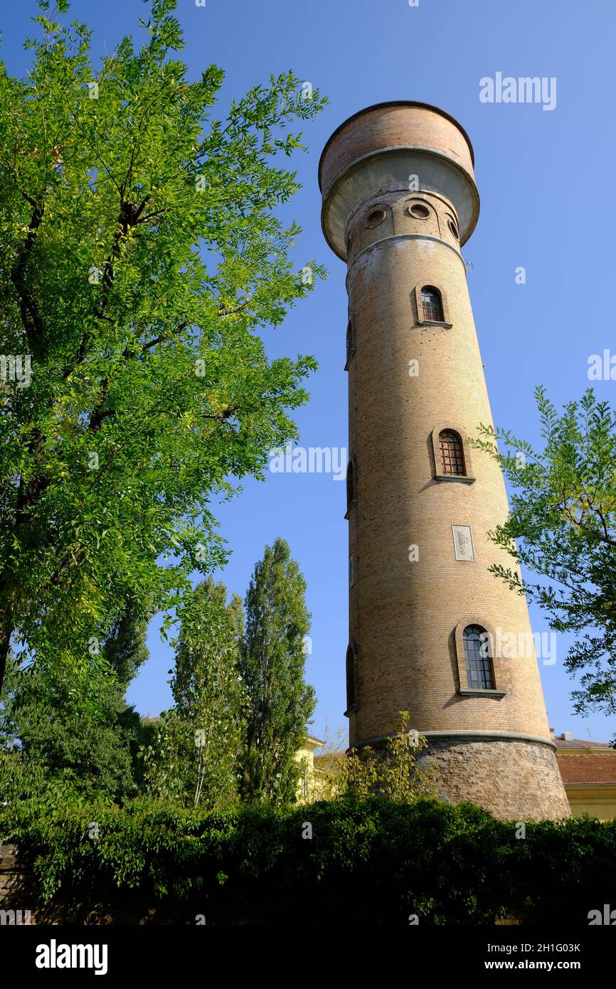 La torre cilindrica in mattoni si innalza nel cielo blu. Torre acquedotto con finestre ad arco, costruita con mattoni di terracotta e circondata da alti alberi di pioppo. Foto Stock