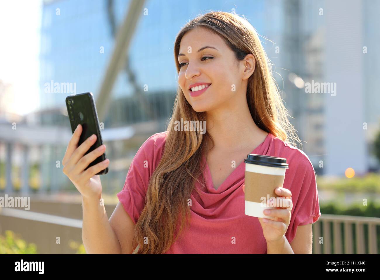Giovane donna brasiliana sorridente con blusa rosa videochiama con smartphone all'aperto, tenendo una tazza di caffè da asporto Foto Stock