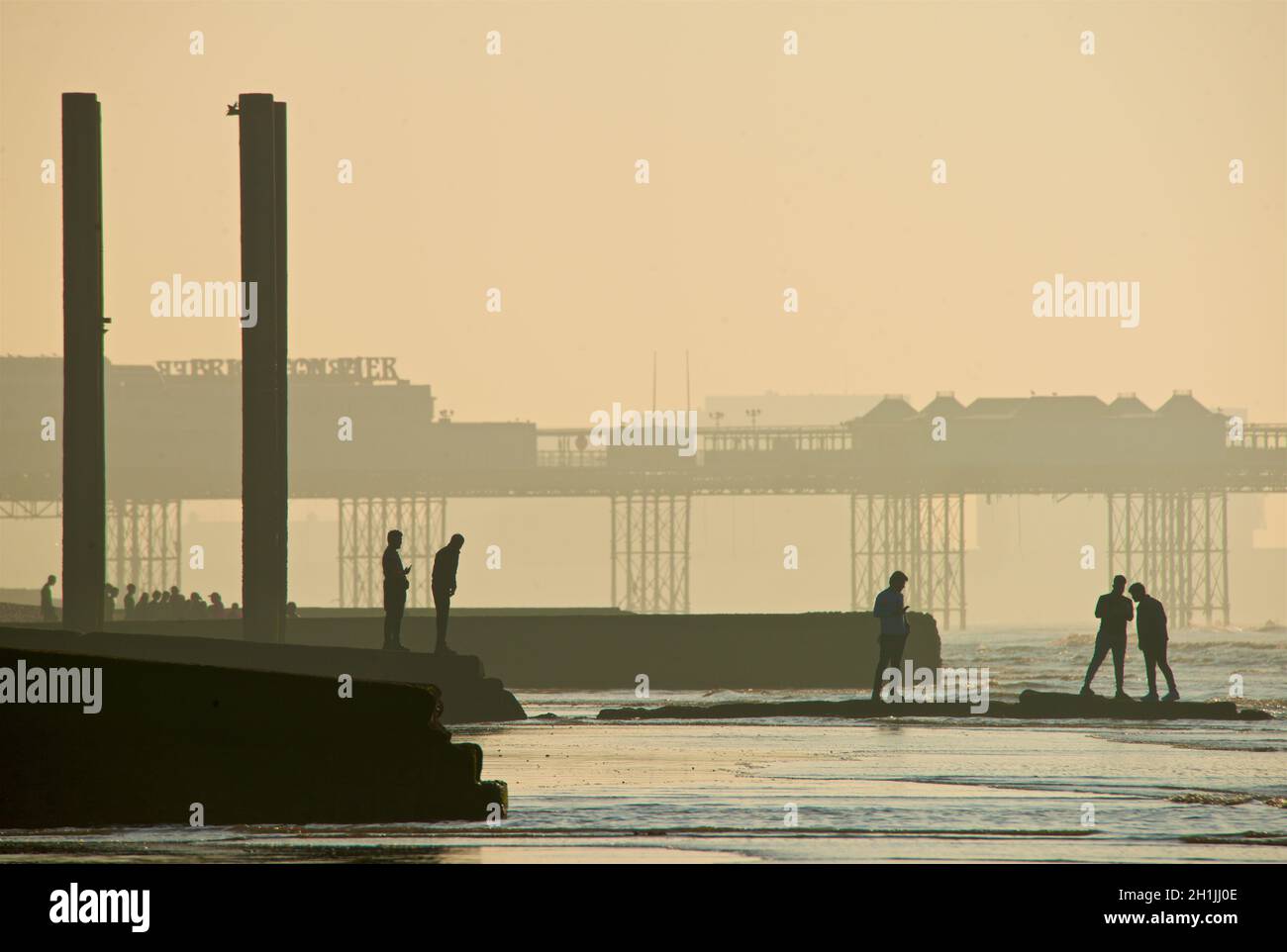 Forme silhoueted di cinque amici su un groyne che conduce al mare a bassa marea. Condivisione selfie. Brighton & Hove, East Sussex, Inghilterra, Regno Unito. Pillars Beach End del vecchio West Pier; Palace Pier dietro. Foto Stock