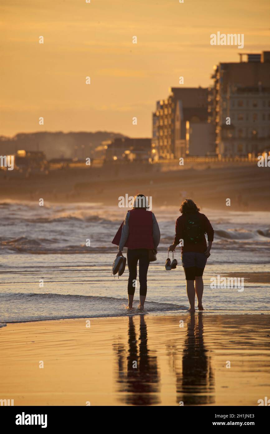 Due donne si addentrano nelle acque poco profonde a bassa marea, camminando verso ovest verso Hove. Tenendo le scarpe nelle mani. Brighton & Hove, East Sussex, Inghilterra, Regno Unito Foto Stock