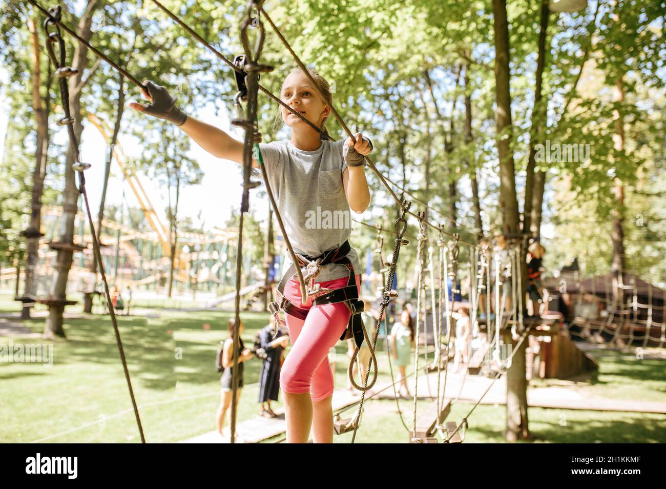 Piccola ragazza sorridente in attrezzatura sale in corda parco, parco giochi. Bambini che si arrampicano su ponte sospeso, avventura sportiva estrema in vacanza, pericolo it Foto Stock